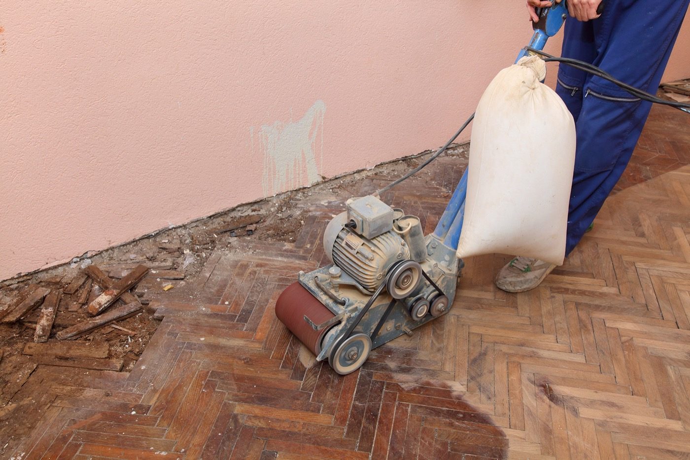 Operaio con levigatrice a nastro e sacco raccoglipolvere su un parquet a spina di pesce rovinato, con listelli staccati lungo la parete